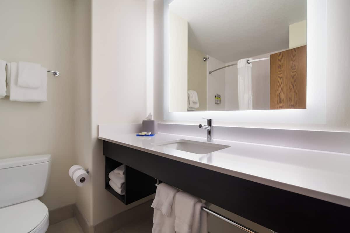 A modern bathroom with a white sink, a mirror, and a toilet. There is a towel rack with towels and a shelf with toiletries.