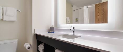 A modern bathroom with a white sink, a mirror, and a toilet. There is a towel rack with towels and a shelf with toiletries.