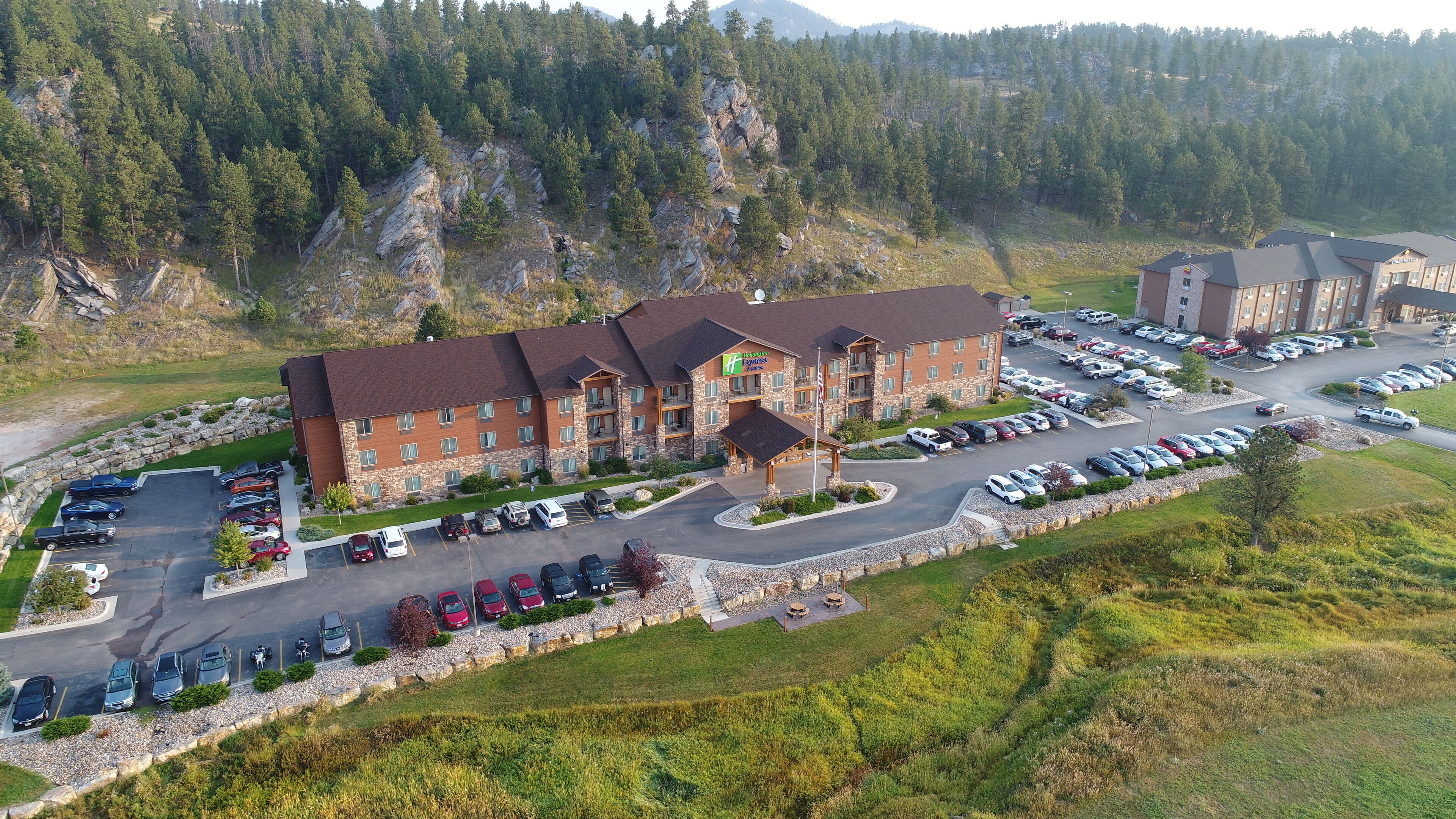 A hotel with a central building, a parking lot, and a mountainous backdrop.