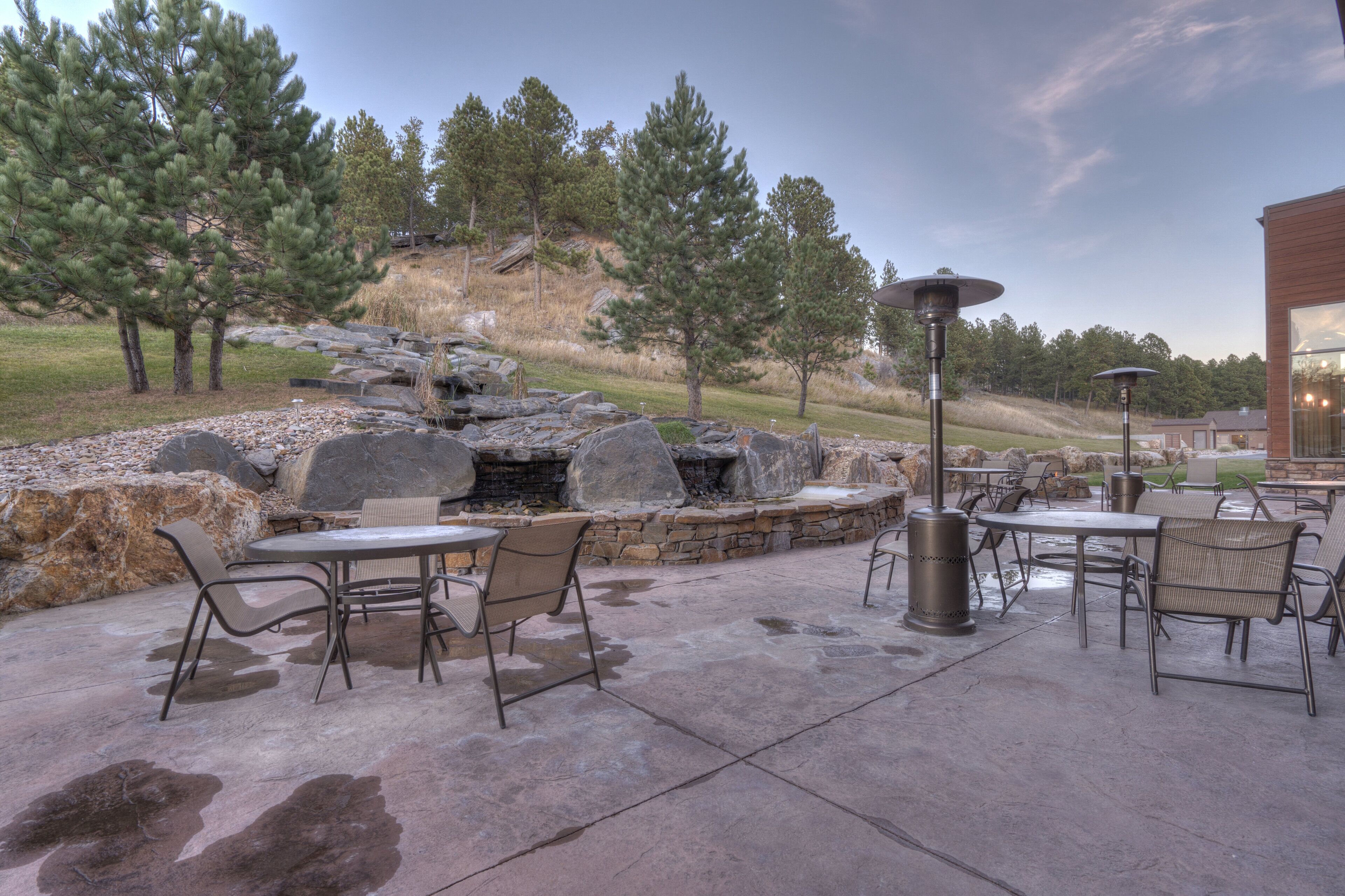 An outdoor patio with stone tables and chairs, a stone waterfall feature, and a view of a hilly landscape with trees.