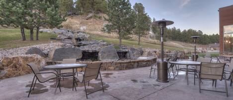 An outdoor patio with stone tables and chairs, a stone waterfall feature, and a view of a hilly landscape with trees.