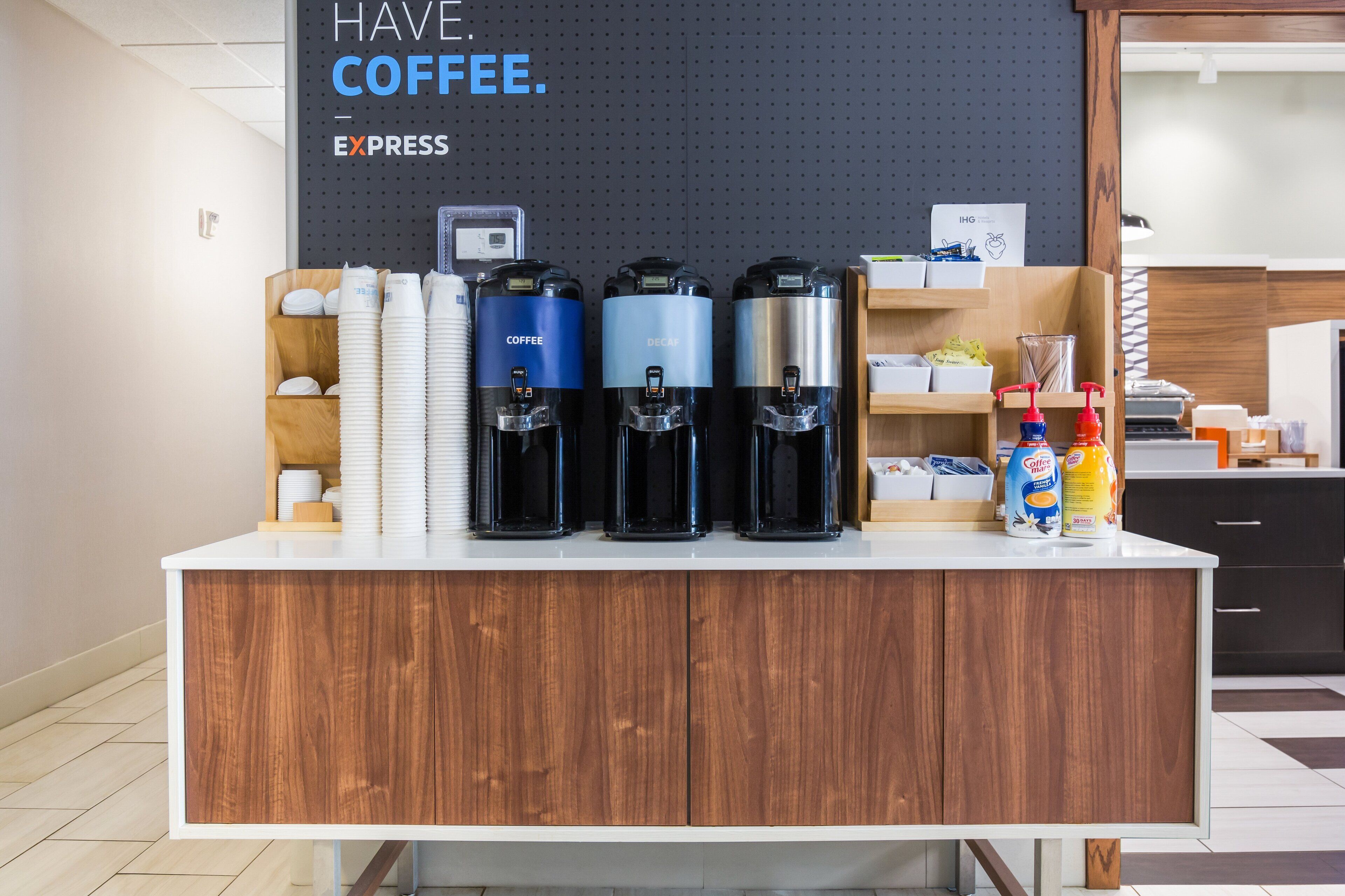 A coffee station with three coffee dispensers, a stack of white cups, and a sign that says "HAVE COFFEE. EXPRESS".
