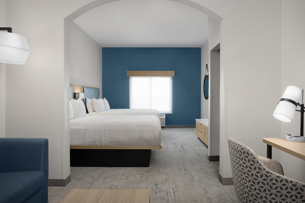 A modern hotel room with a large bed, a desk, and a chair. There is a window with blinds, a lamp, and a decorative wall piece.