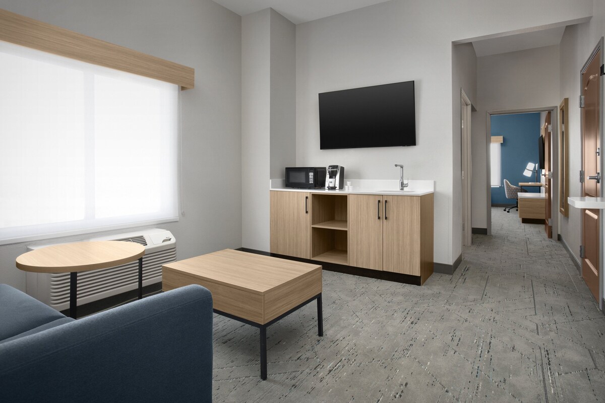 A modern hotel room with a flat-screen TV, a kitchenette, a sofa, and a dining table.