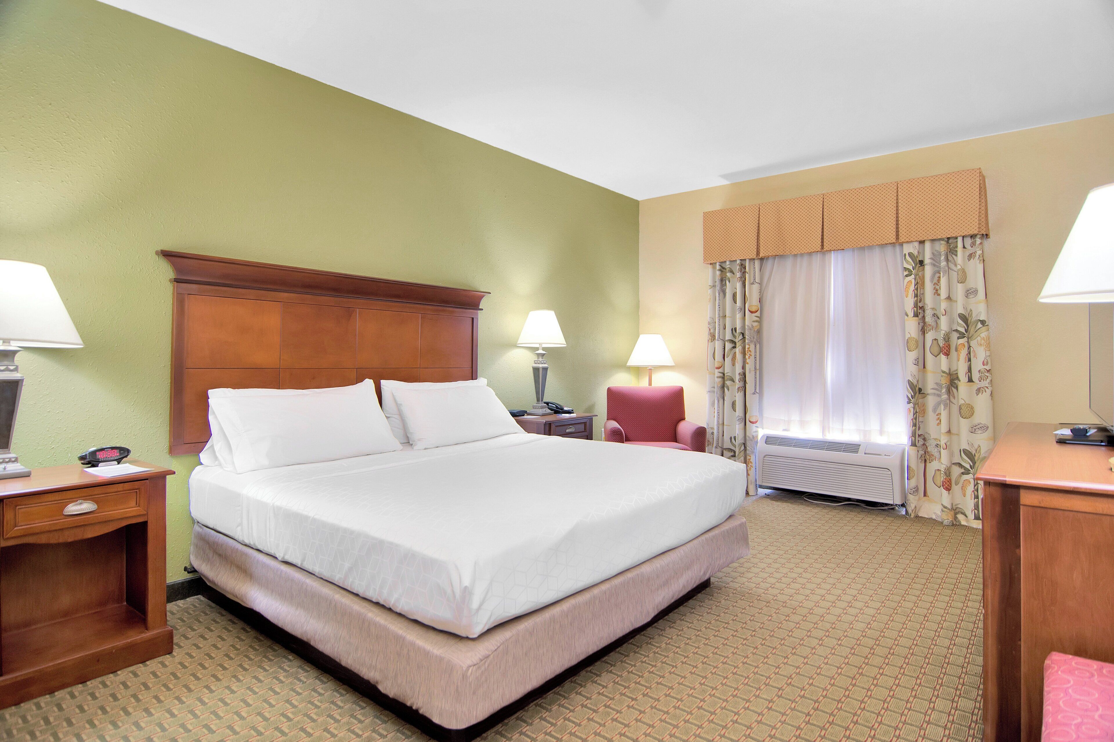 Une chambre d’hôtel avec un grand lit, deux tables de chevet, un bureau, une chaise, une fenêtre avec des rideaux et un climatiseur fixé au mur.