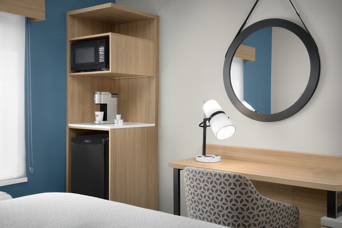 A modern hotel room with a bed, a desk with a lamp, a microwave, a coffee maker, and a dishwasher.