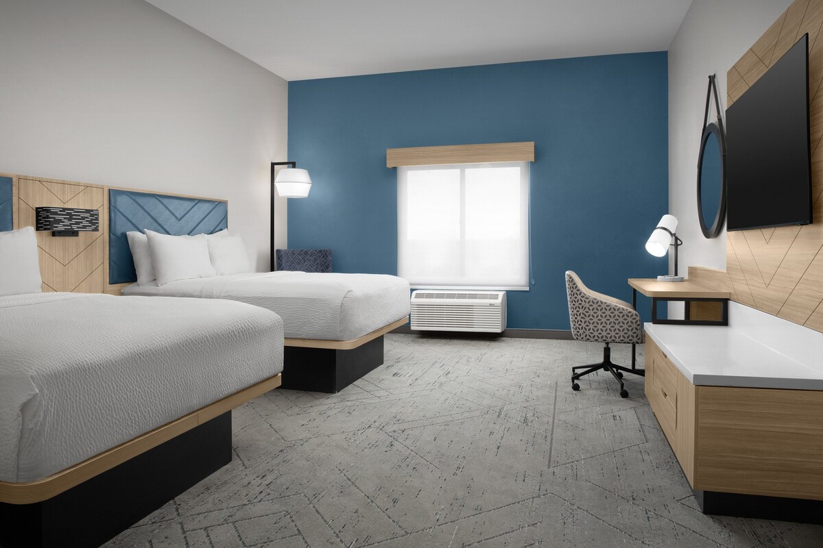 A modern hotel room with two beds, a desk, a chair, a large window, and a flat-screen TV.