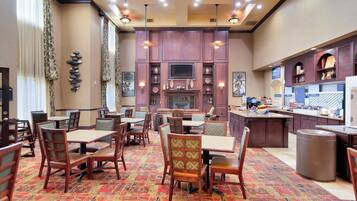 A hotel dining area with multiple tables and chairs, a kitchen area with a buffet, and a patterned carpet.