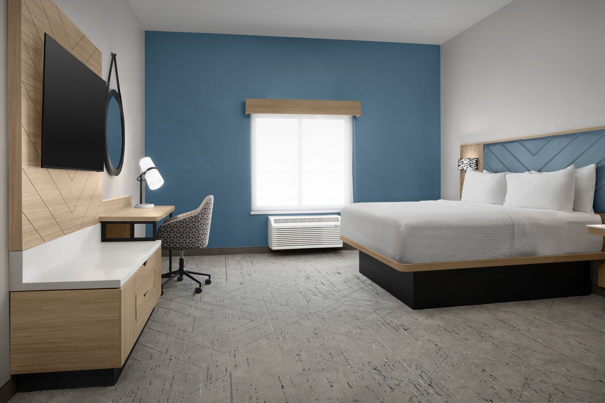 A modern hotel room with a large bed, a desk with a chair, a flat-screen TV, and a window with blinds.