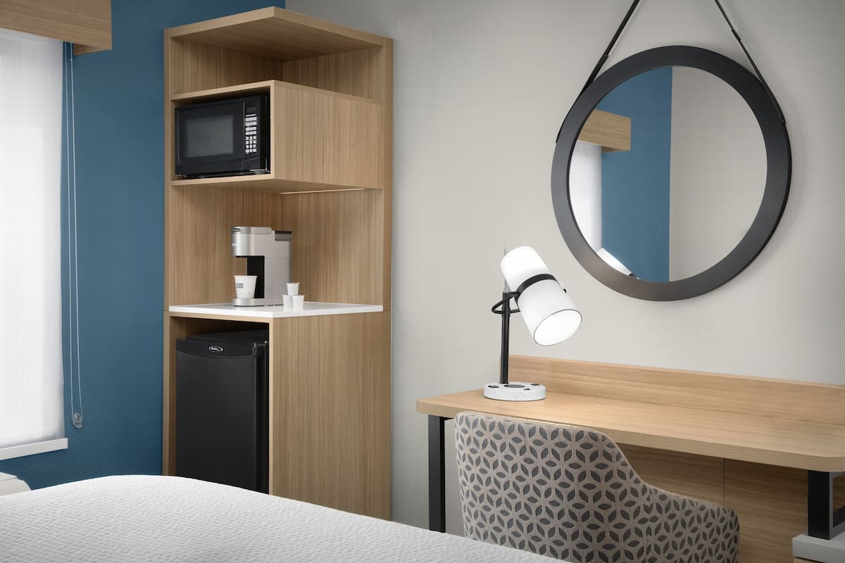 Una habitación de hotel moderna con una cama, un escritorio con lámpara, microondas, cafetera y lavavajillas.
