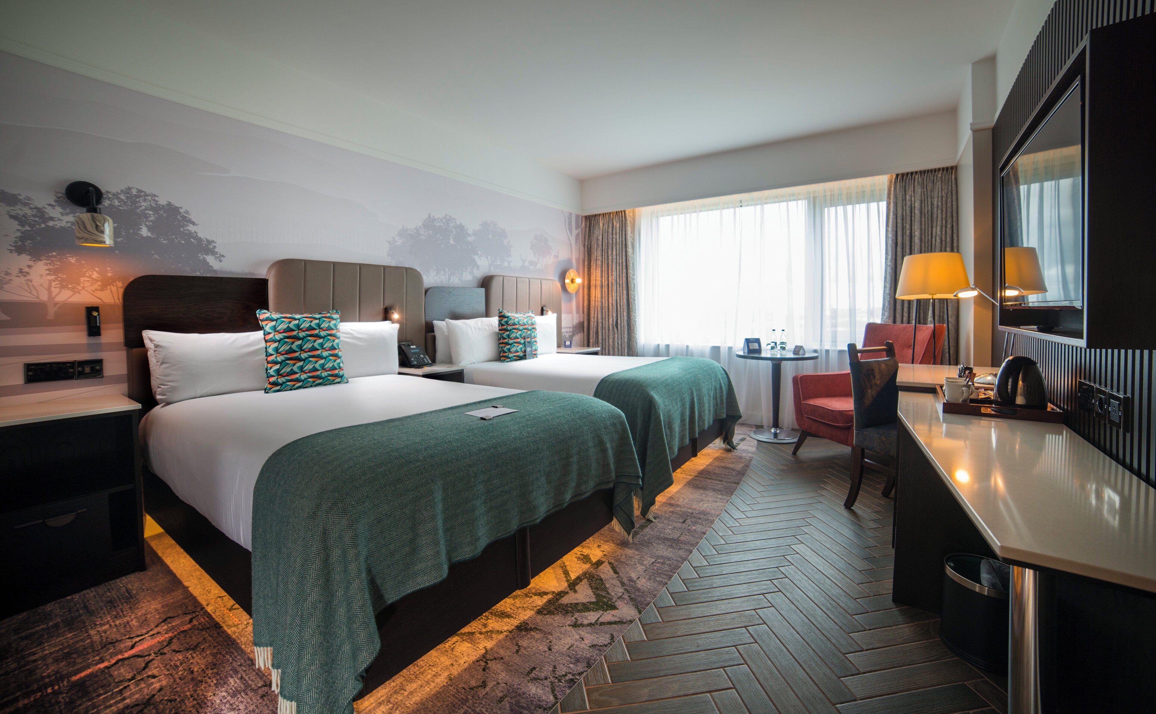 A hotel room with two beds, a desk, a chair, a mirror, and a window with curtains.