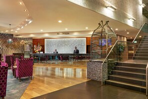 Un espace d’accueil d’hôtel comprenant un comptoir de réception, un grand mur orné de citations inspirantes et un escalier doté d’une rampe en métal.