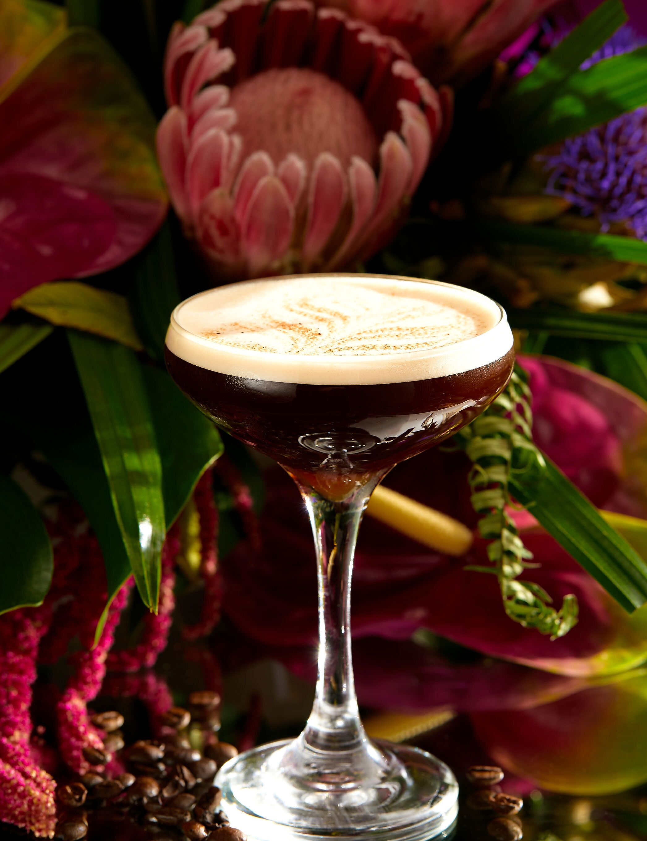 A cocktail in a stemmed glass with a creamy foam on top, surrounded by vibrant flowers and greenery.