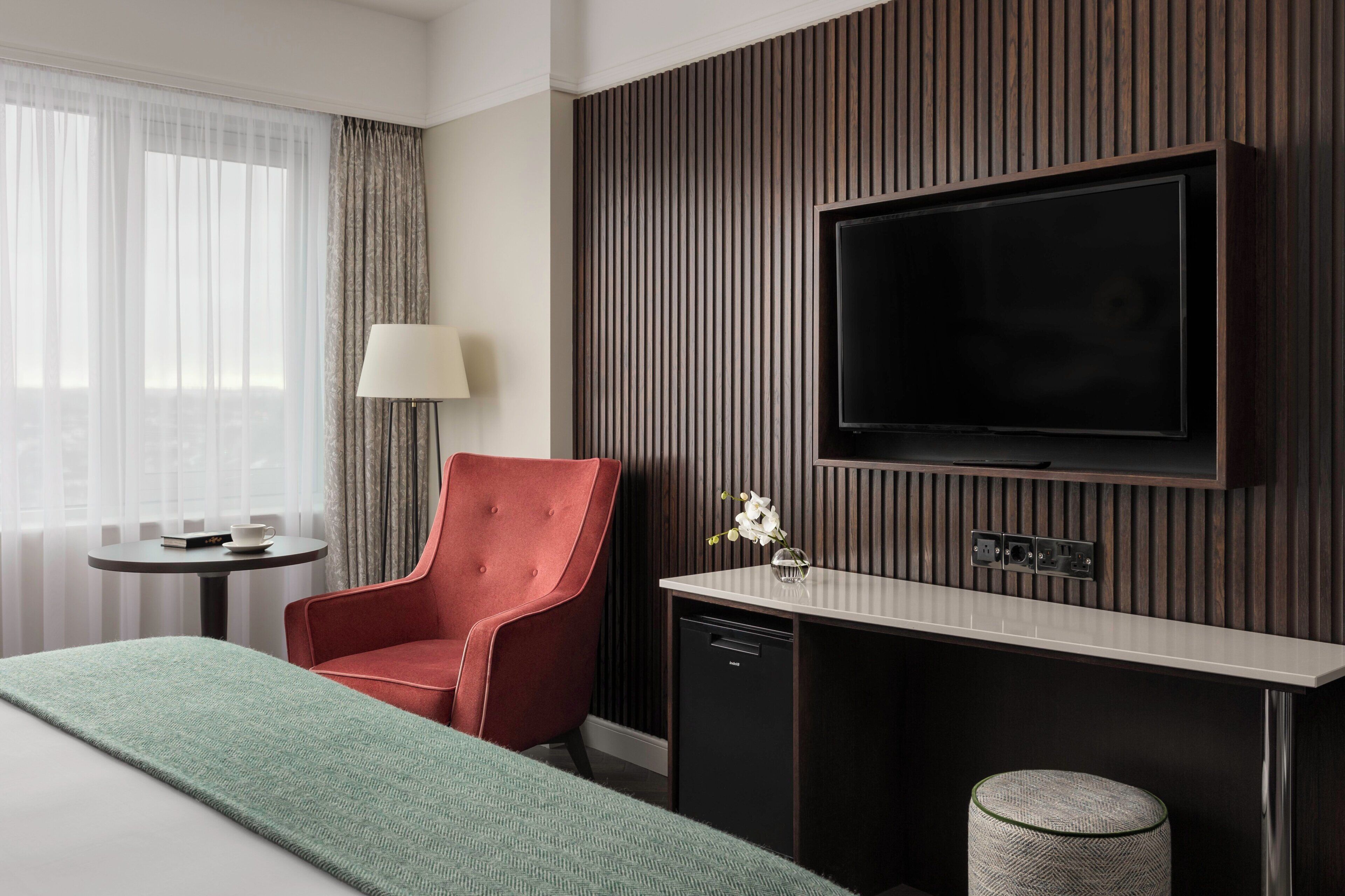 Une chambre d’hôtel avec un lit, un fauteuil rouge, une petite table avec une lampe, un téléviseur fixé au mur et une prise électrique murale.
