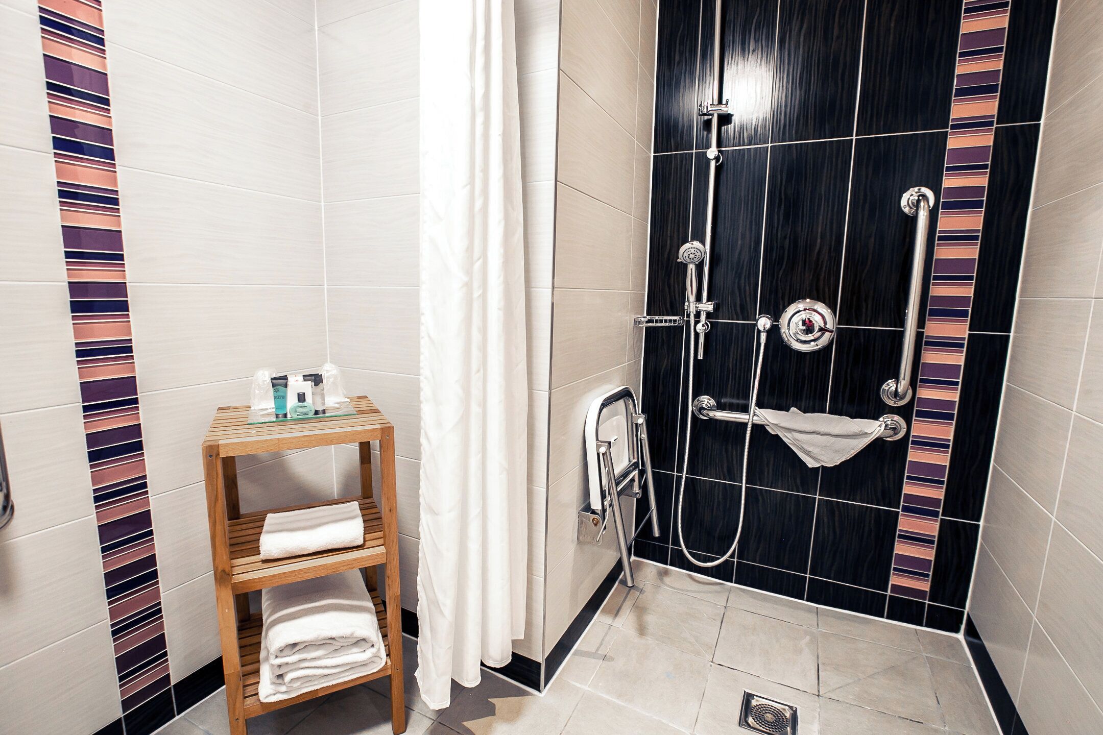 A bathroom with a shower, a wooden shelf with towels, and a tiled wall.