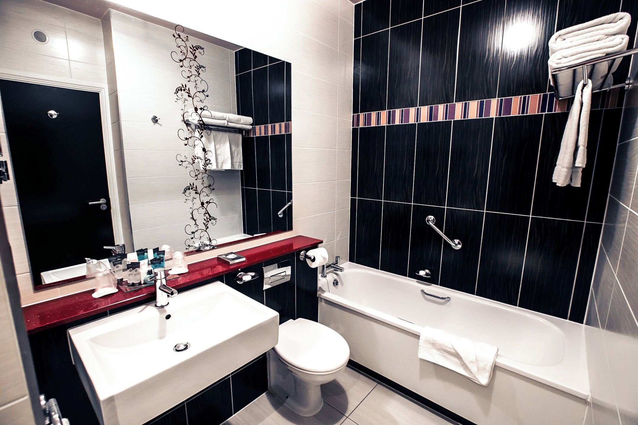 Et moderne badeværelse med en hvid vask, toilet og badekar. Et spejl er monteret over vasken, og der er en håndklædestang over badekarret.