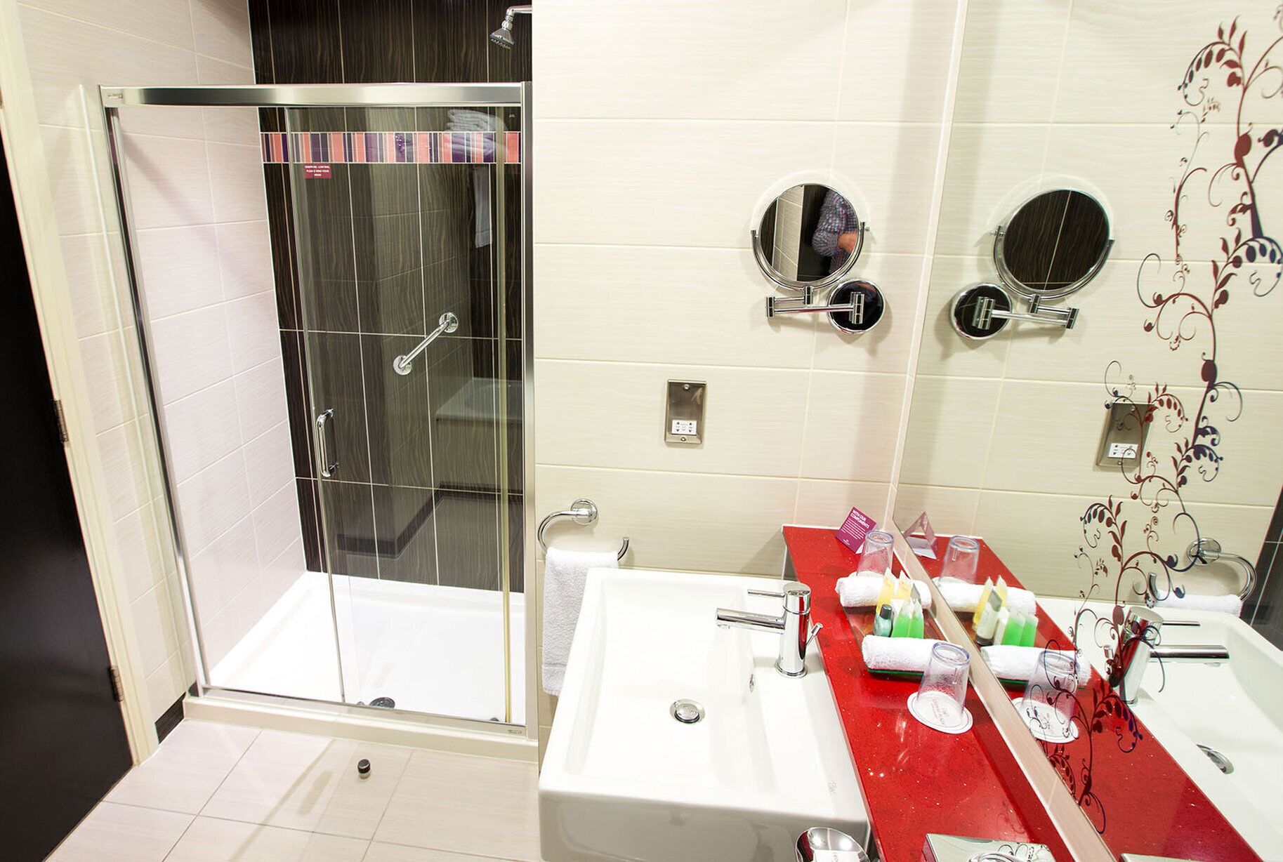 A bathroom with a glass-enclosed shower, a sink with a red countertop, and two wall-mounted mirrors.
