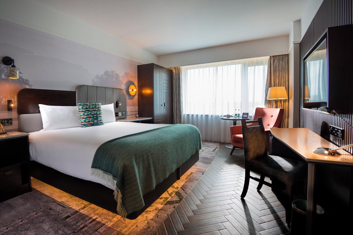 Habitación de hotel con una cama grande, un escritorio, una silla, una mesita, una lámpara y una ventana con cortinas.