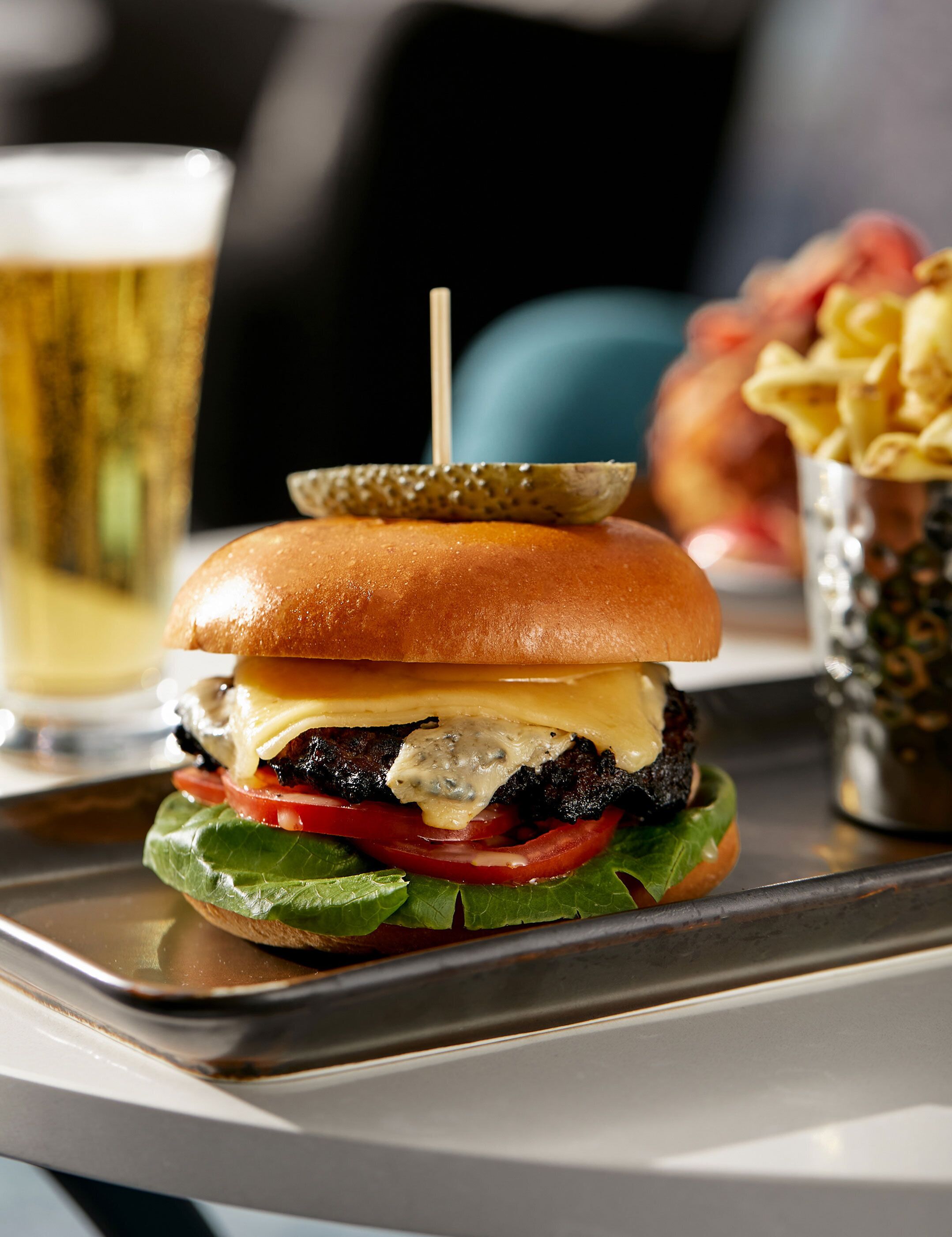 A burger with a toasted bun, cheese, lettuce, tomato, and a grilled patty, accompanied by a glass of beer and a side of fries.