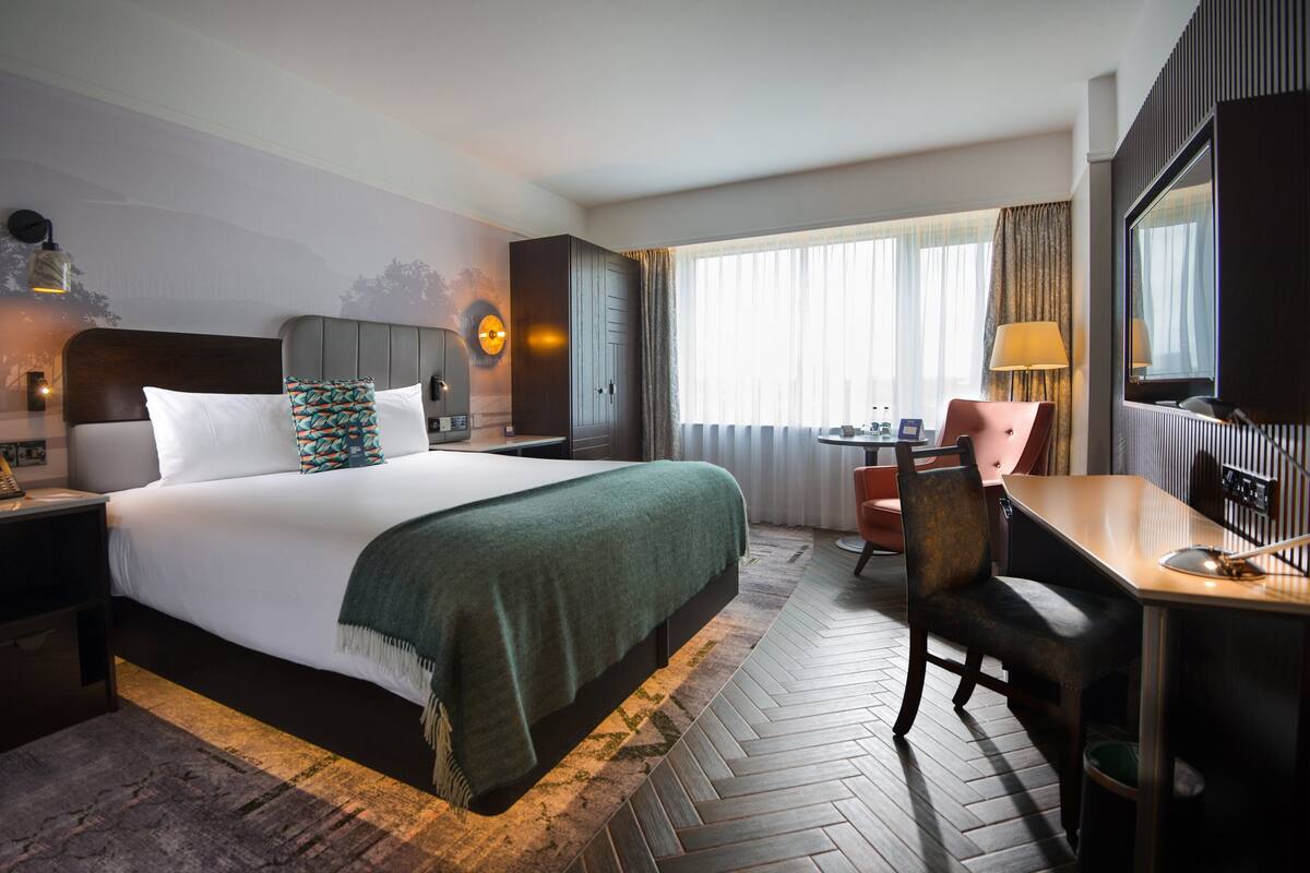 Habitación de hotel con una cama grande, un escritorio, una silla, una mesita, una lámpara y una ventana con cortinas.
