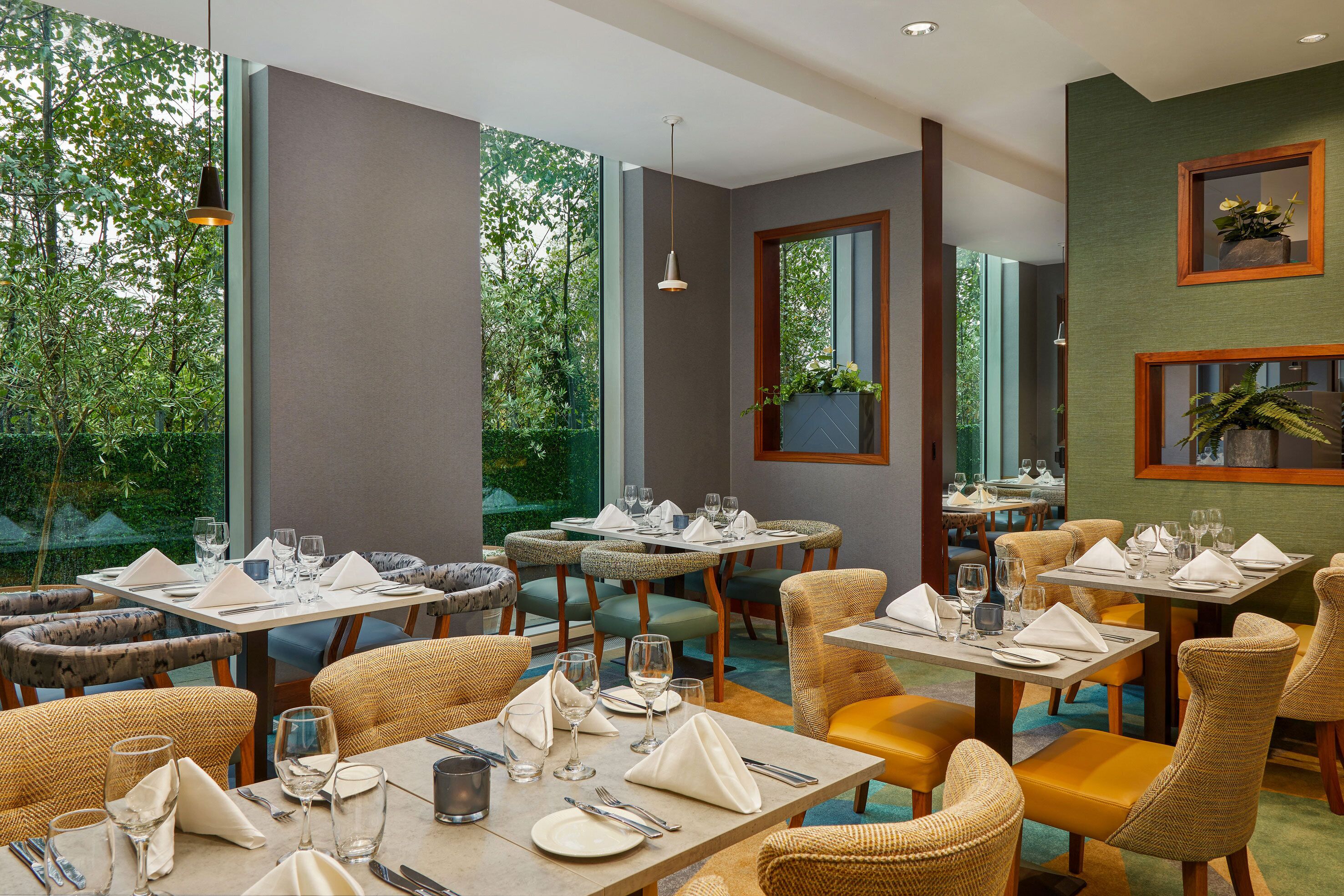 Un restaurant avec des tables dressées pour le service, des chaises et de grandes fenêtres donnant sur un paysage verdoyant.