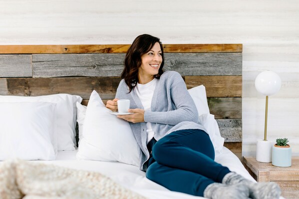 A person sitting on a bed with white pillows, holding a cup, in a room with a wooden headboard and a nightstand with a lamp and a small plant. - Glacier View Lodge (Jasper)