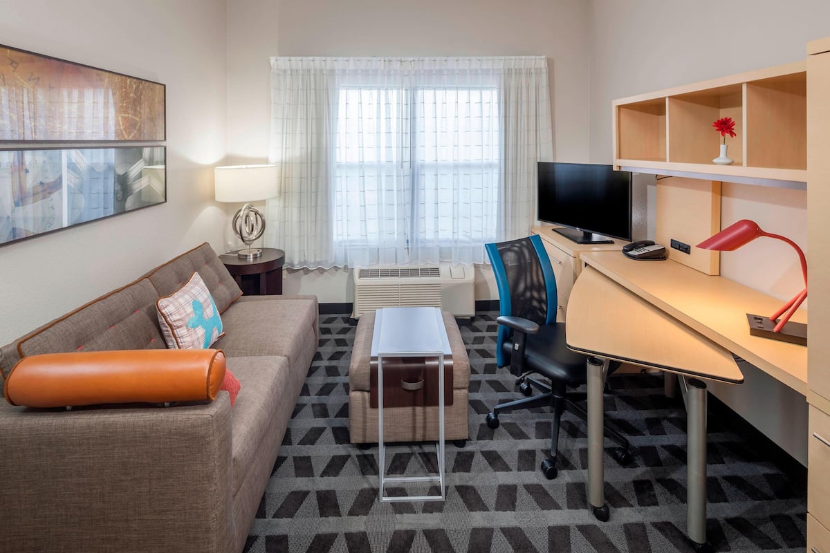 A hotel room with a sofa, a desk, a chair, a television, a lamp, and a window with curtains.