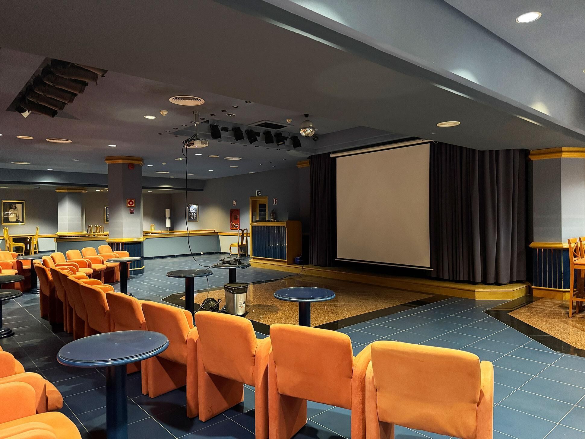 Una sala conferenze con file di sedie arancioni, uno schermo grande e un palco con tende.