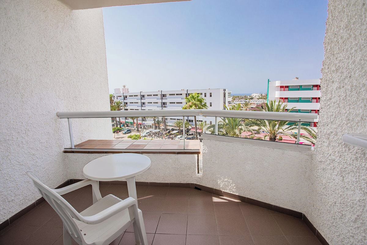 A balcony with a white table and chairs, offering a view of buildings and palm trees.