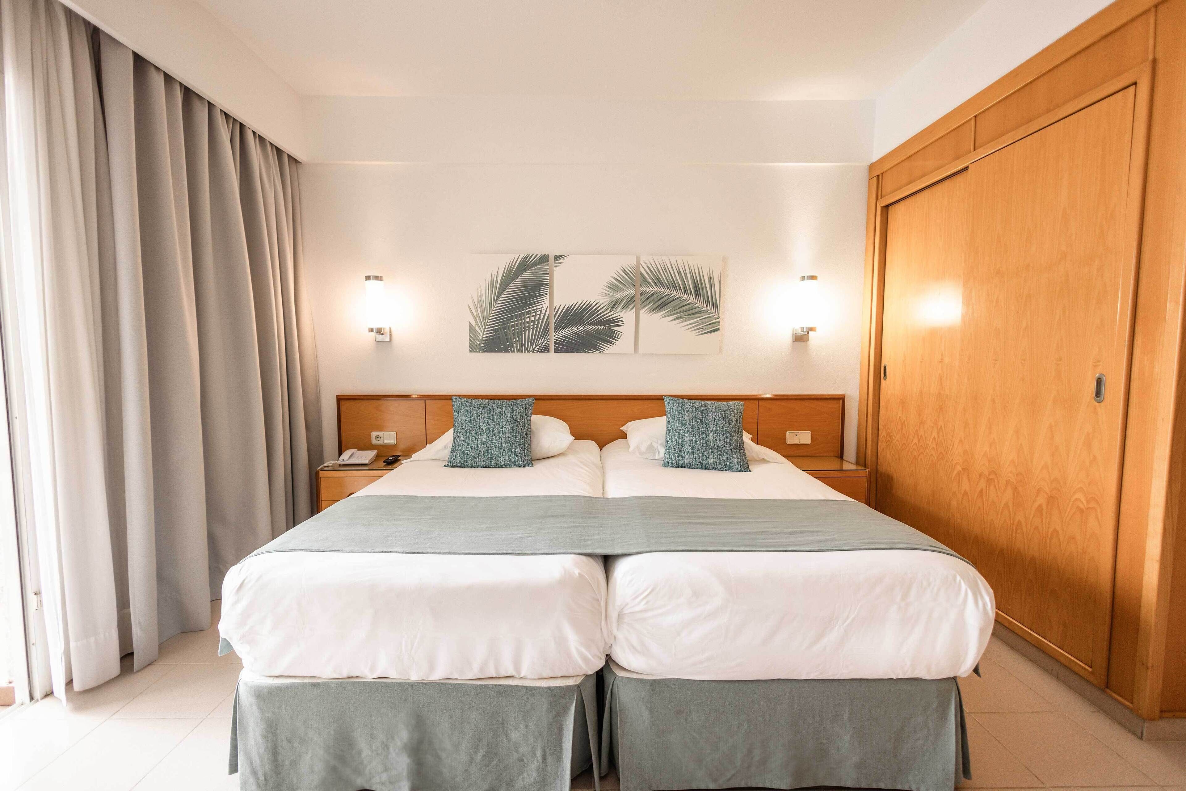 Una cama doble con dos almohadas, una mesita de noche, una lámpara fijada a la pared y un armario de madera grande.