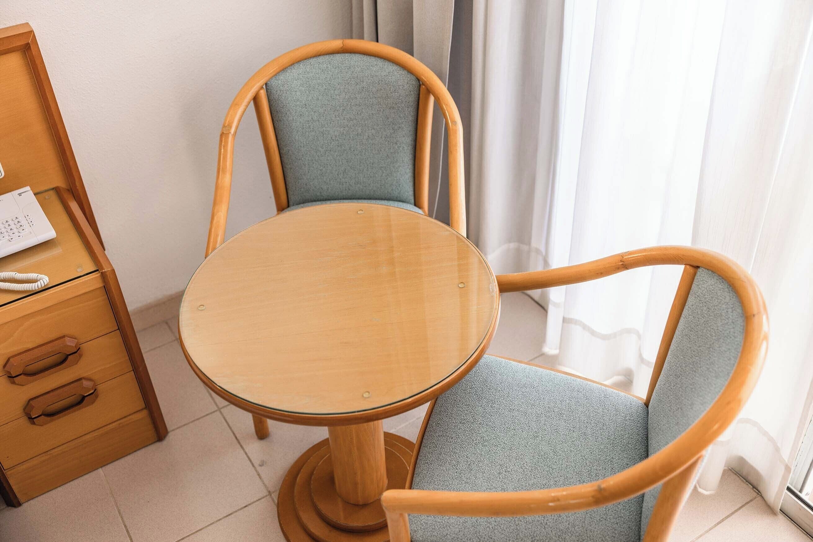 A small wooden table with a glass top, a blue upholstered chair, and a wooden drawer unit with a telephone.