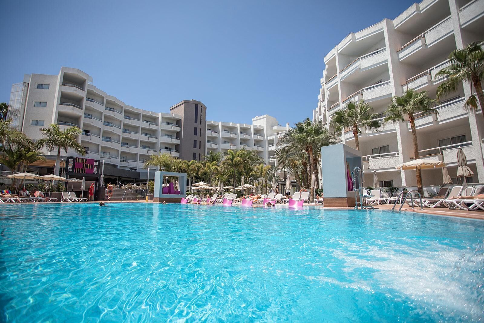 A hotel pool area with multiple levels of balconies, palm trees, and sun loungers.