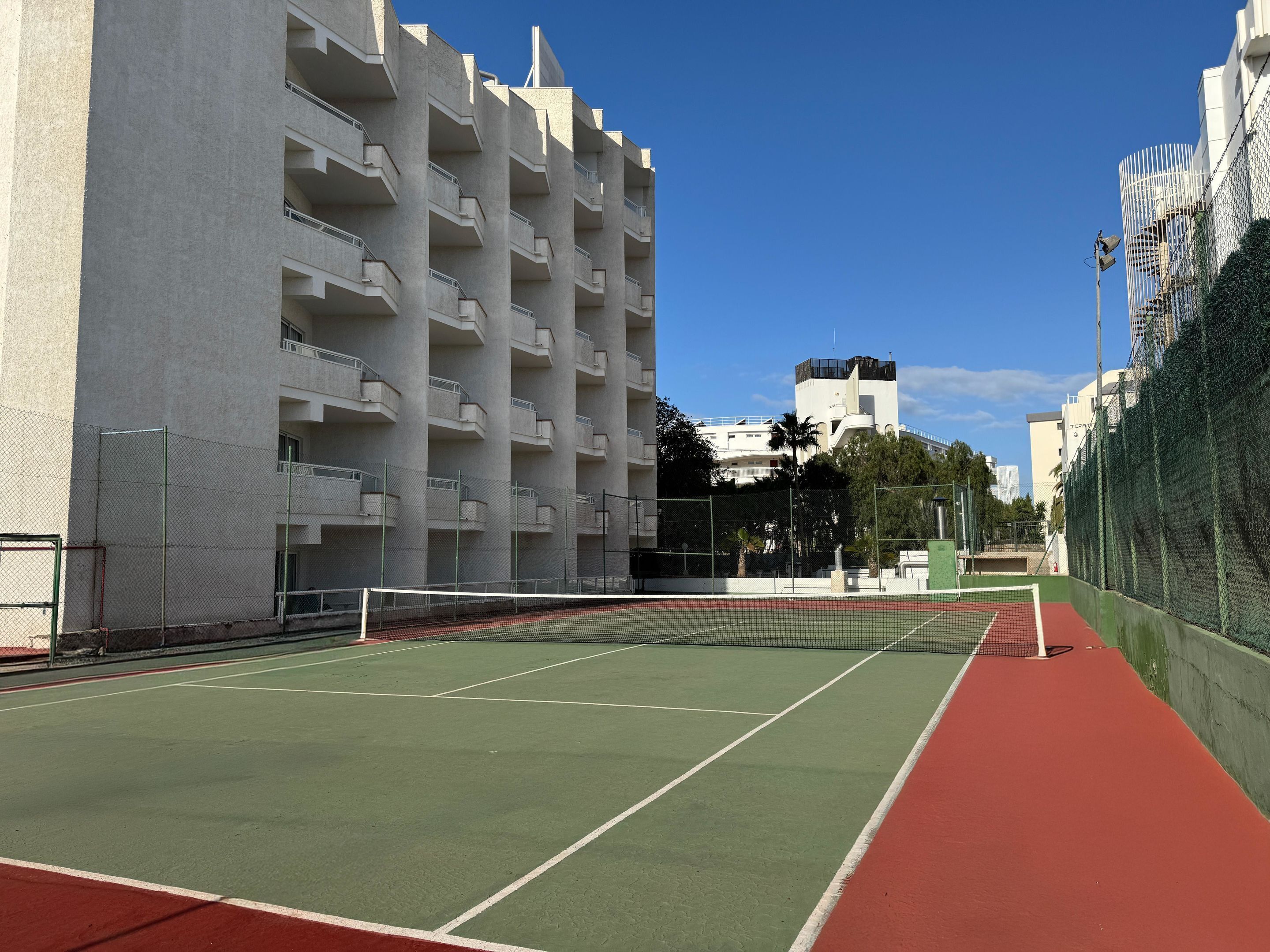 A tennis court with a net, surrounded by a chain-link fence and adjacent to a multi-story building with balconies.