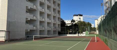 A tennis court with a net, surrounded by a chain-link fence and adjacent to a multi-story building with balconies.