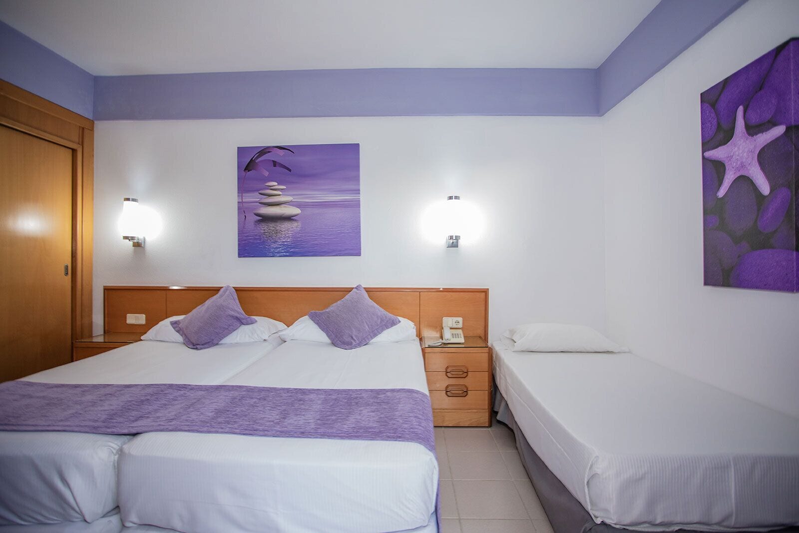 Hotelkamer met een groot bed, een kleiner bed, een nachtkastje, een telefoon en een schilderij van een strandgezicht.