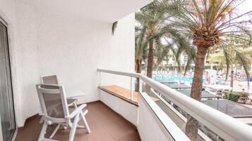 Un balcón con una silla y una mesa blancos, con vista a una zona de piscina con palmeras y edificios.