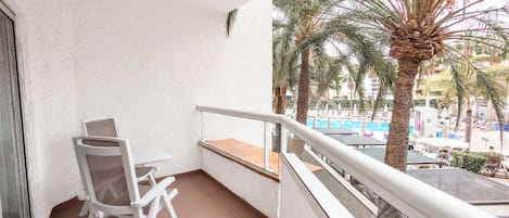 Un balcón con una silla y una mesa blancos, con vista a una zona de piscina con palmeras y edificios.