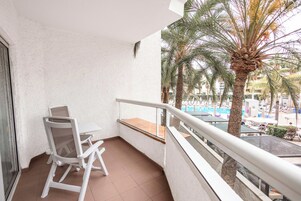 Un balcón con una silla y una mesa blancos, con vista a una zona de piscina con palmeras y edificios.