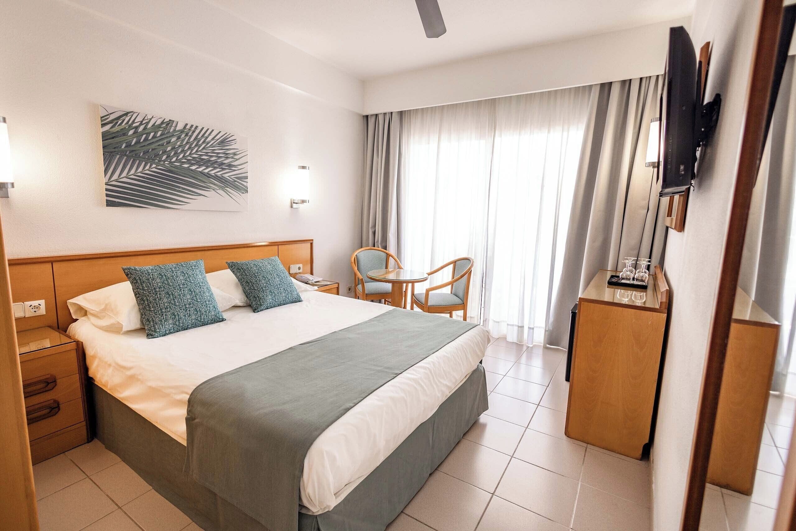 A hotel room with a large bed, a desk with a chair, a TV mounted on the wall, and a window with curtains.
