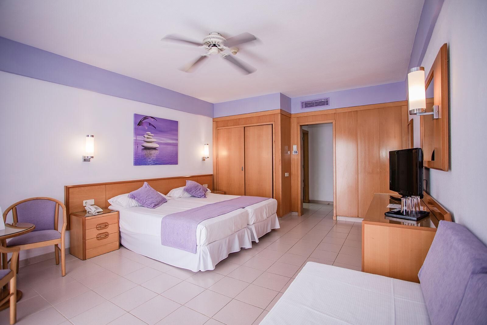 Une chambre d’hôtel comprenant un lit, un bureau avec une télévision, une chaise, une lampe de bureau et un ventilateur de plafond.
