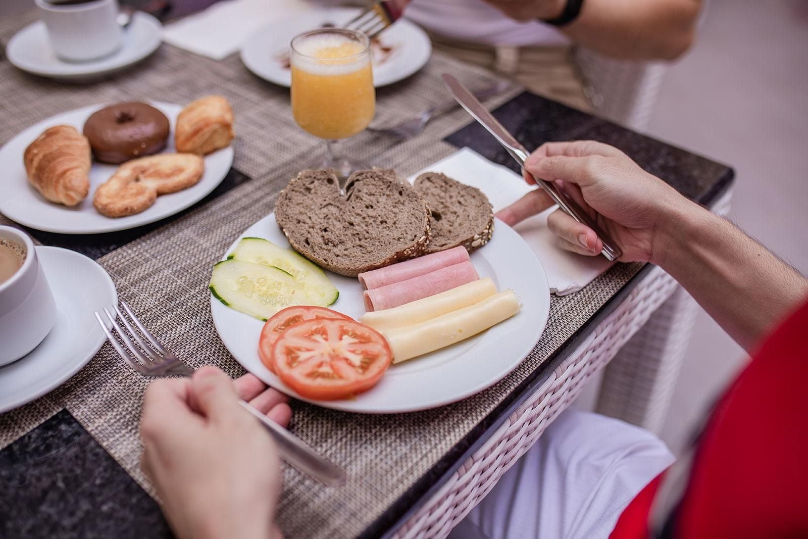 Desayuno con croissants, pan, queso, jamón, tomate, pepino y un vaso de jugo de naranja sobre una mesa.