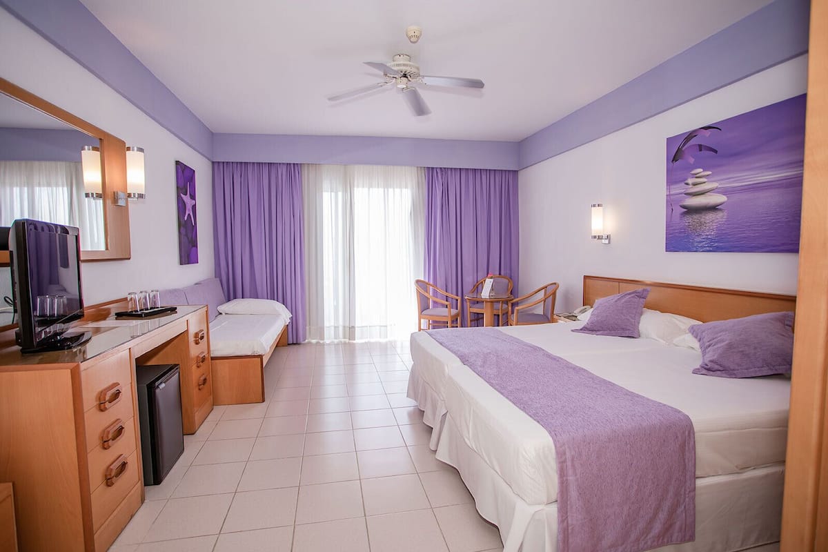 A hotel room with a bed, a desk with a television, a chair, and a window with purple curtains.