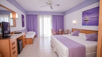 Quarto de hotel com uma cama, uma escrivaninha com televisão, uma cadeira e uma janela com cortinas roxas.