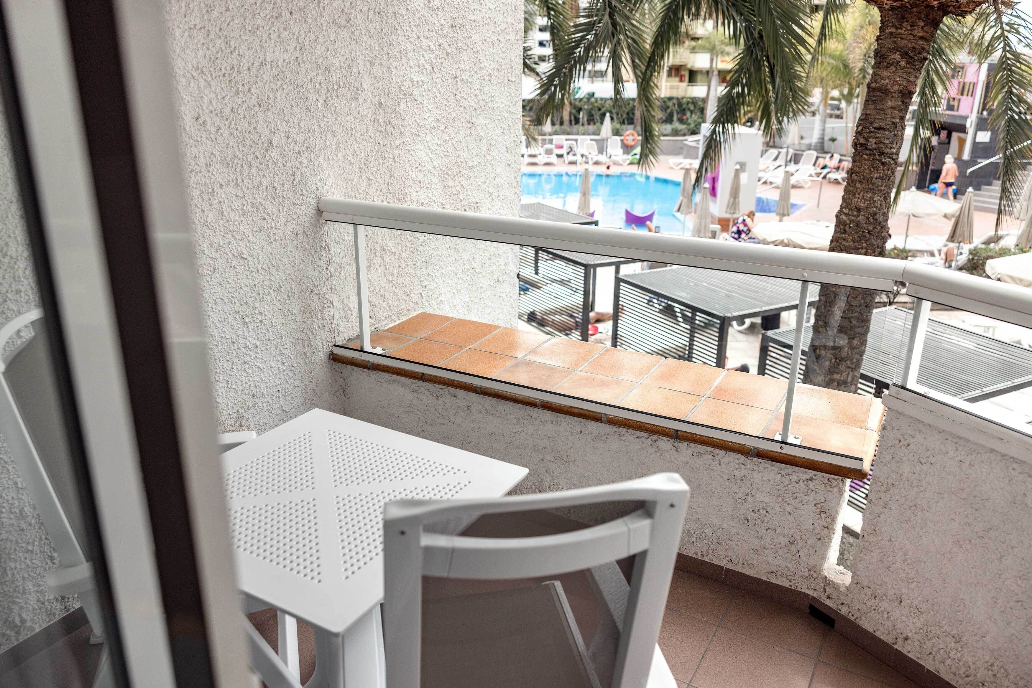 Un balcon avec une chaise et une table blanches, donnant sur un espace piscine équipé de chaises longues et de parasols.