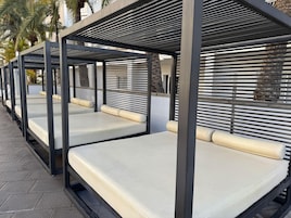Filas de zonas de estar al aire libre con estructuras metálicas y cojines color beige, frente a un fondo de palmeras y un edificio.