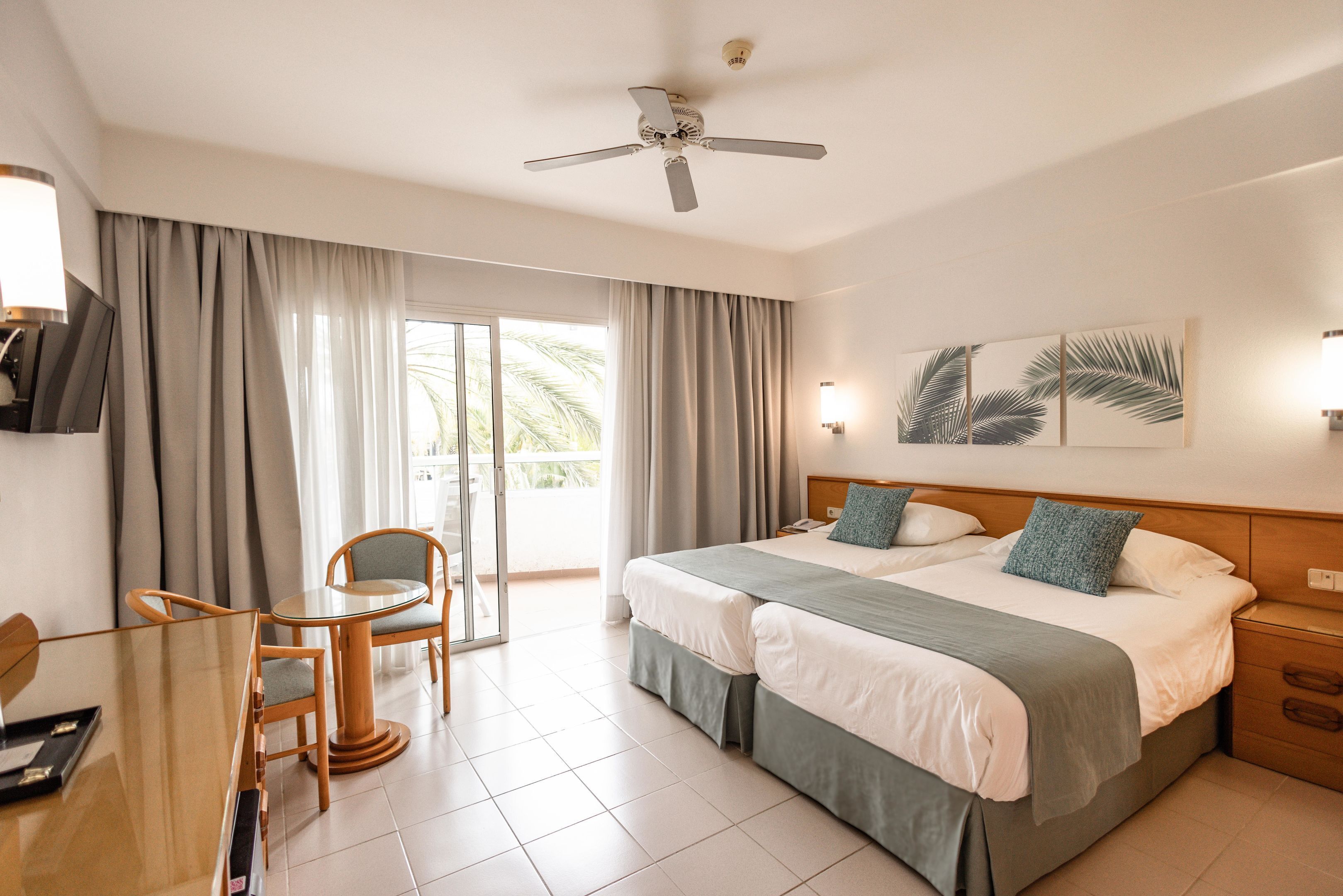 A hotel room with a bed, a desk, chairs, a ceiling fan, and a large window with curtains.