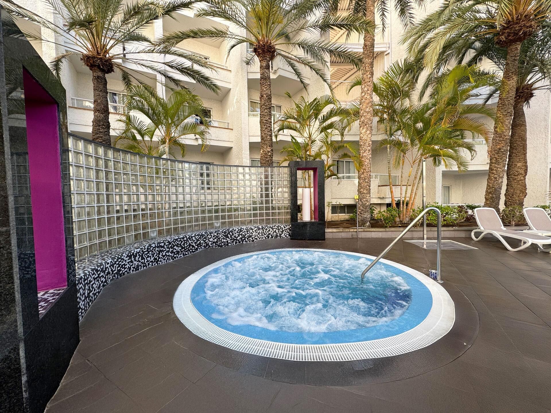 Ein Whirlpool, umgeben von Palmen und Liegestühlen, mit einer gefliesten Wand und einer violetten Säule.
