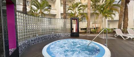 A hot tub surrounded by palm trees and lounge chairs, with a tiled wall and a purple pillar.