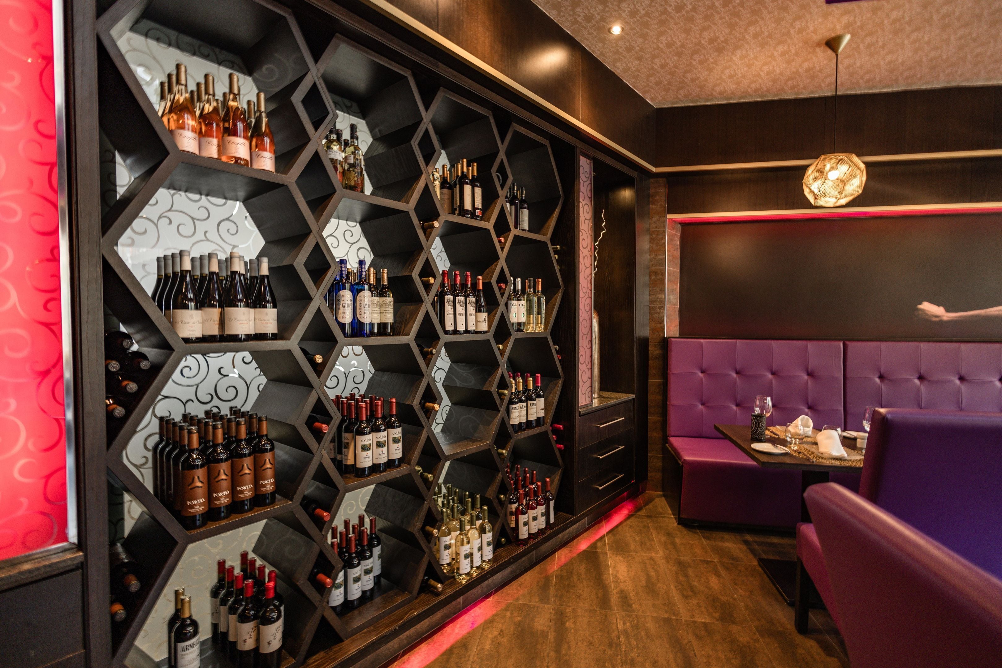 Een bar met hexagonaal open vakken vol flessen, een paars zitgedeelte en een moderne plafondlamp.