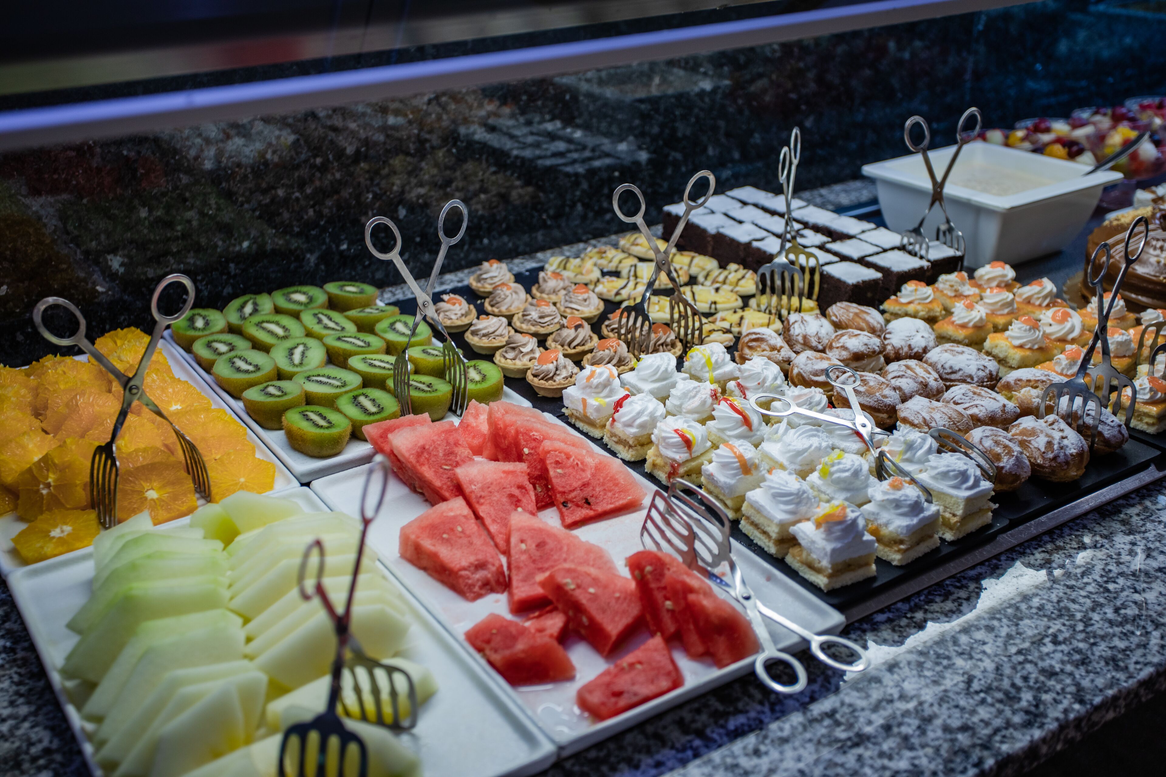 Eine Auswahl an Desserts und Früchten, darunter Wassermelone, Kiwi und Gebäck, ansprechend auf Tellern mit Besteck arrangiert.